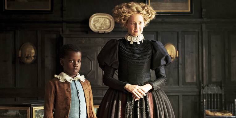 Ein Schwarzer Junge und eine weiße Frau in einem Kleid mit Reifrock stehen in einem Raum vor einer dunklen Wand. (Schwarz groß geschrieben)
